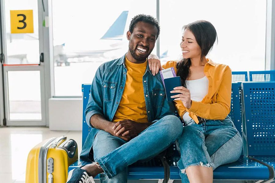 Travel Insurance - Smiling Young Couple Sitting in the Departure Lounge with Baggage and Tickets in the Airport with Airplane behind in the Distance