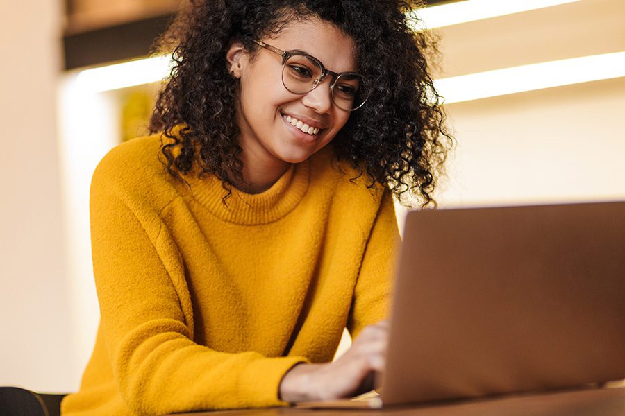 Blog - Woman in Yellow Sweater Smiling at Open Laptop While Typing and Sitting at a Table